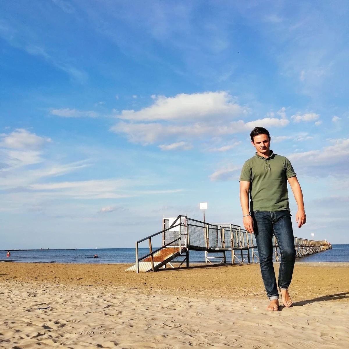 Filippo walking barefoot on the beach in Rimini, the pier behind him