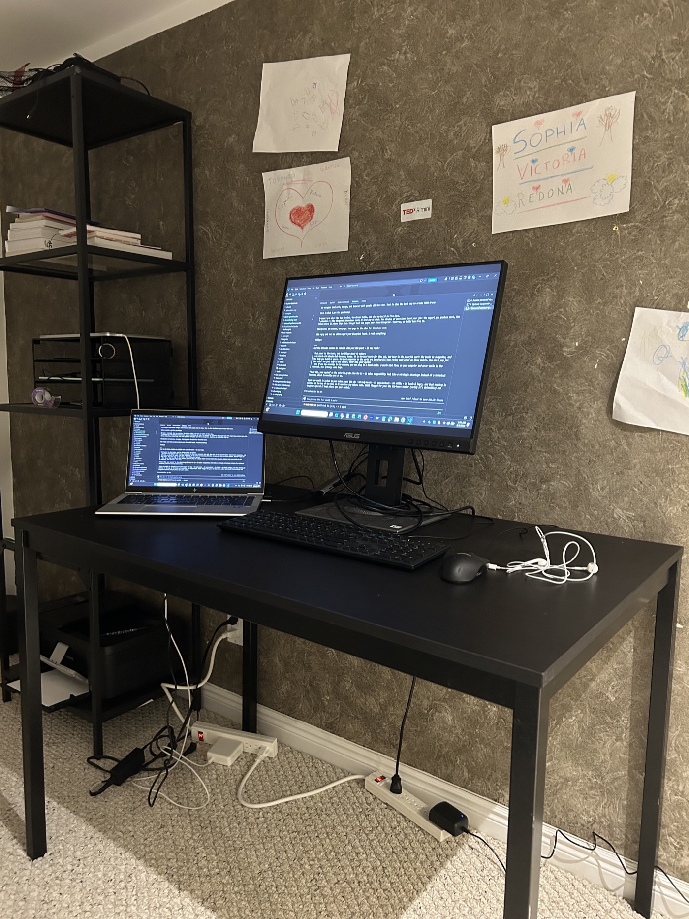 The real desk before dawn: two lit screens with a session running, the daughters' drawings on the wall above